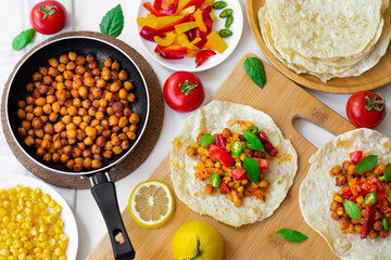 Vegan tortilla tacos with chickpeas, bell peppers, corn, tomatoes, basil leaves and green chilli. On the background are ingredients, lemons, chickpeas in a small pan