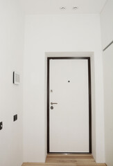 White entrance hall, entryway in a renovated apartment with black and white armored door, a peephole, door handle and door lock, smoke detector and home automation smart home control panel.