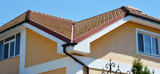 A close-up on a red clay tiled roof valley, fascia and rain gutter system with a downspout of a...