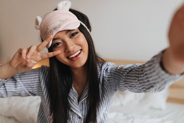 Girl in cute sleeping mask shows sign of peace and makes selfie in bedroom