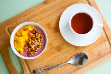 Healthy summer breakfast of a pink smoothie bowl made with frozen banana and raspberry garnished with granola and fresh orange. In the background is black tea