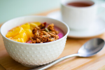 Healthy summer breakfast of a pink smoothie bowl made with frozen banana and raspberry garnished with granola and fresh orange. In the background is black tea