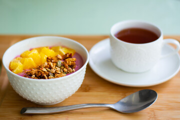 Healthy summer breakfast of a pink smoothie bowl made with frozen banana and raspberry garnished with granola and fresh orange. In the background is black tea