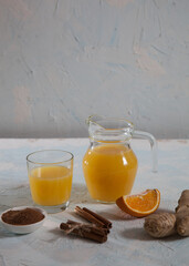Orange juice with ginger in a glass jug and in a glass, cinnamon sticks on a light background. Health. Copy spaes.