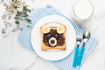 Food art bear toast for kids with chocolate nut spread, fruits and glass of milk. Top view on blue...