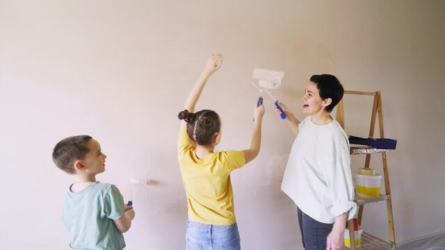 joyful mum with little son and daughter colours new apartment wall and clap rollers with happiness standing near wooden ladder with tools close backside view