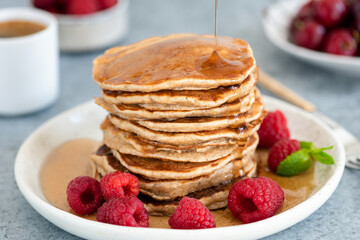 Syrup pouring on stack of whole wheat pancakes served with fresh raspberries on a plate. Closeup...