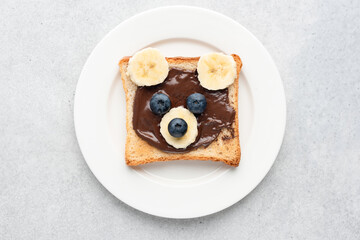 Kids breakfast or lunch toast shaped as teddy bear, food art. White sandwich bread with chocolate nut spread and fruits, children's food menu. Isolated on white plate on grey concrete background