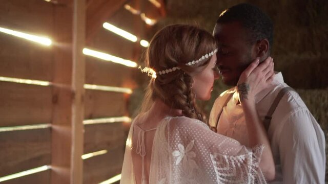 Happy Caucasian bride and African groom dressed in boho style are hugging in a barn in the sun. Close up.