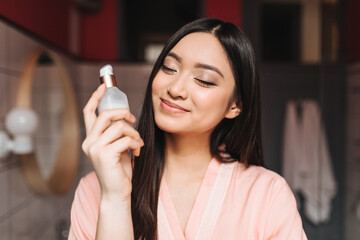 Optimistic Asian girl smiles with closed eyes and holds jar of cream