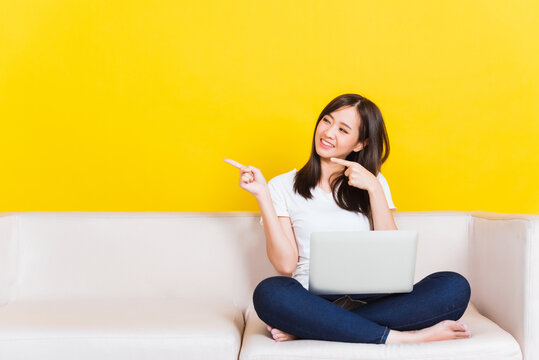 Portrait Asian Of Happy Beautiful Young Woman Work From Home She Sitting On Sofa Using Laptop Computer In House Living Room Pointing Finger To Side Studio Shot Isolated On Yellow Background