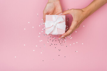 Women hands holding a gift or gift box decorated with confetti on a pink pastel table top view