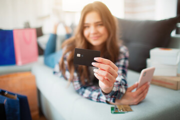 online shopping. A young woman lies on a sofa at home and shows a close-up of credit card planks to the camera.