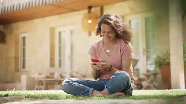 Young woman sits on green grass on lawn in country house and chatting smartphone. High quality 4k footage