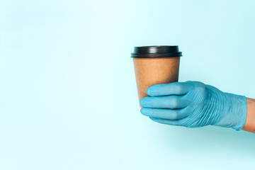 Hand in medical gloves carrying paper cup of takeaway coffee on blue background. Banner with copy space. Contactless delivery service during quarantine coronavirus pandemic. Take away only concept