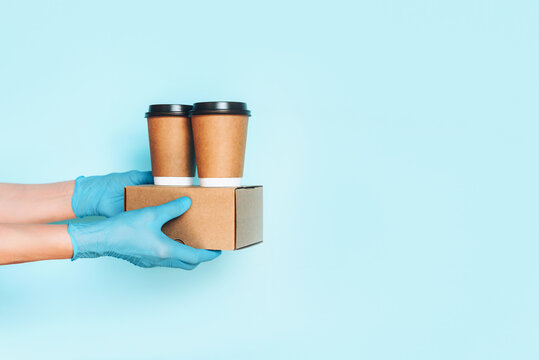 Hand In Medical Gloves Carrying Paper Coffee Cup Coffee, Lunch Box On Blue Background. Banner, Copy Space. Contactless Delivery Service During Quarantine Coronavirus Pandemic. Take Away Only Concept