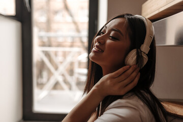 Close-up shot of Asian woman wearing headphones and white T-shirt against window