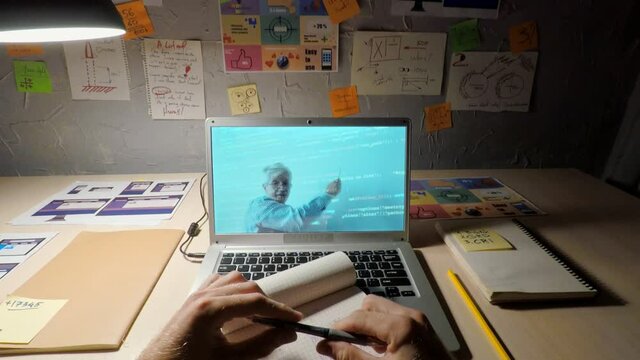 POV shaky shot of unrecognizable male student watching online computer programming lesson with senior teacher on laptop screen and taking notes in notepad sitting at desk at home