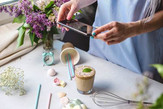 A woman is taking photos on a mobile phone camera of a whipped morning Dalgona coffee. White background, pastel colors and lilac flowers