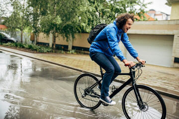 Fototapeta premium Horizontal outdoor image of handsome man cycling on his bike down the street next to the house. Caucasian male courier with curly hair delivers parcel cycling with a bicycle in the city in a rainy day