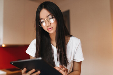 Pensive Asian girl looks into screen of tablet. Brunette woman in glasses posing in kitchen