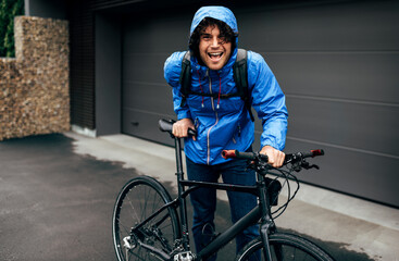 Smiling handsome man walking with his bike after bicycling down the street on a rainy day next to the fence's house. Cheerful male courier with curly hair delivers parcel cycling with a bicycle.