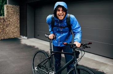 Foto op Plexiglas Smiling handsome man walking with his bike after bicycling down the street on a rainy day next to the fence's house. Cheerful male courier with curly hair delivers parcel cycling with a bicycle. © iuricazac