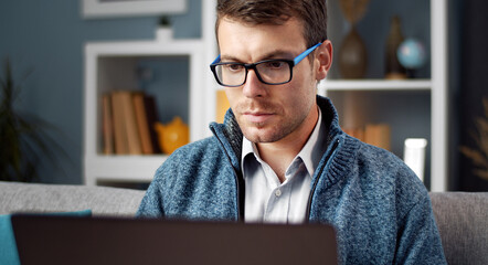 Focused man in eyewear and casual clothes using laptop looking at screen sitting on sofa in...