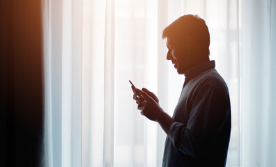 Silhouette of man looking at mobile phone standing sideways to curtained window, half-body shot