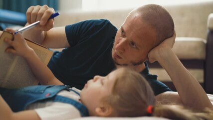 CU, cam tracking: little girl student and her bald unshaven dad lie on floor and read book, hold pen and pencil, discuss, prompt and help her, smile, closeup