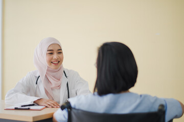 Young Asian woman Muslim doctor  giving advice discussion and check up to elderly patient sitting in Wheelchair at hospital which smiling and felling happy. Medicine and health care safe concept.