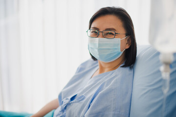 Elderly Asian patient woman smiling and felling happy under mask sitting in bed at hospital. Medicine and health care safe concept.