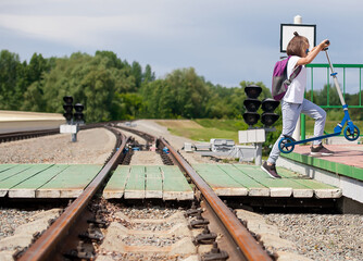 Naklejka premium Crossing the railway tracks made of wood across the railway tracks in the daytime. Schoolgirl carrying scooter and crossing road. Pedestrian passing a crosswalk. Safety concept for road users