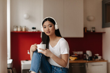 Brown-eyed girl in white T-shirt and massive headphones looks at camera, posing with cup on background of kitchen