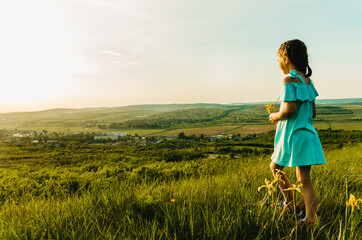 Portrait of little caucasian girl in blue dress admiring the sunset over rural landscape on a hill