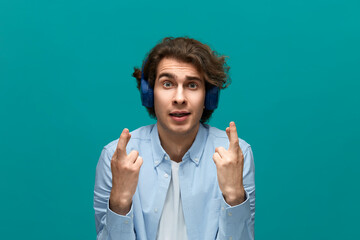 I hope. Portrait of a young beautiful man wearing white t-shirt and blue shirt in blue headphones holding his fingers crossed and looking at camera with pleading