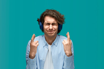 Hard expectation. Portrait of a young beautiful man wearing white t-shirt and blue shirt in blue headphones holding his fingers crossed and squinting with pleading