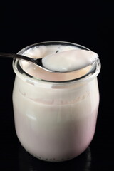 Spoon with yogurt on open small glass jar at black background. M