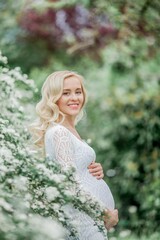 Naklejka premium Young beautiful pregnant woman in a lace white dress walks in a flowering park. Portrait of a beautiful pregnant blonde. Spring.