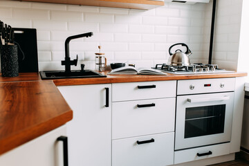 Fototapeta premium Big cook book on the wooden table. Metallic teapot on the gas oven. White kitchen interior. Wooden complete kitchen with gas oven.