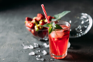 A refreshing summer drink made from strawberries and mint, juice, soda with ice cubes and berry slices, in a beautiful glass with water droplets on a wooden board, dark background