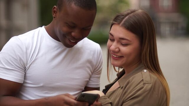 Interracial couple having fun with smartphone in the city street. Young lifestyle concept. A black man and a Caucasian woman laugh and watch photos in a smartphone. Mixed race couple date
