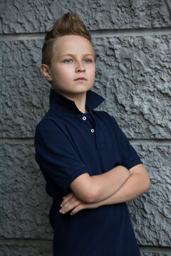 A handsome boy with a fashion haircut is standing by the wall in the street. He is 13 years old. The eyes of the boy are different colors, blue and green.