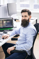 Cheerful senior programmer developer working in IT office, sitting at desk and coding, working on a project in software development company or startup. High quality image.
