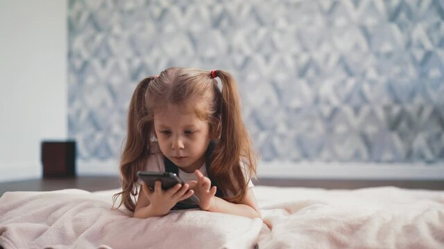 close up of cute little girl with blond long hair braided in pigtails in blue dress lies on floor in room under sunlight, child studies smartphone and video conference