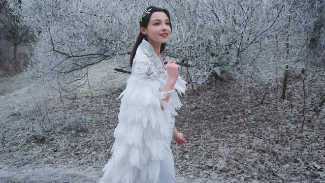 beautiful fantasy woman walking in carnival costume snow queen. White creative cape cloak, silver tiara, diadem, bird white feathers dress. Image greek goddess angel. winter nature snowy trees, forest