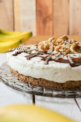 frisch gebackene Torte mit Bananen, Sahne, Schokosoße, Karamellsoße und Haselnussboden auf Tisch aus Holz in der Küche zum Kaffeetrinken