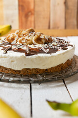 frisch gebackene Torte mit Bananen, Sahne, Schokosoße, Karamellsoße und Haselnussboden auf Tisch aus Holz in der Küche zum Kaffeetrinken