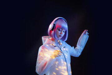 Beautiful woman with purple hair in futuristic costume over dark background. Blue and violet neon light. Portrait of young girl in modern headphones listening music. Free space for text.