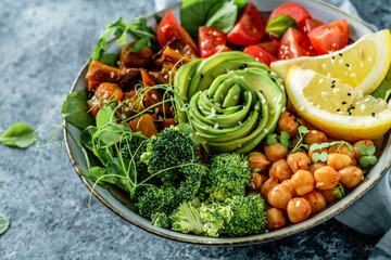 Buddha bowl salad with baked sweet potatoes, chickpeas, broccoli, tomatoes, greens, avocado, pea sprouts on light blue background with napkin. Healthy vegan food, clean eating, dieting, closeup
